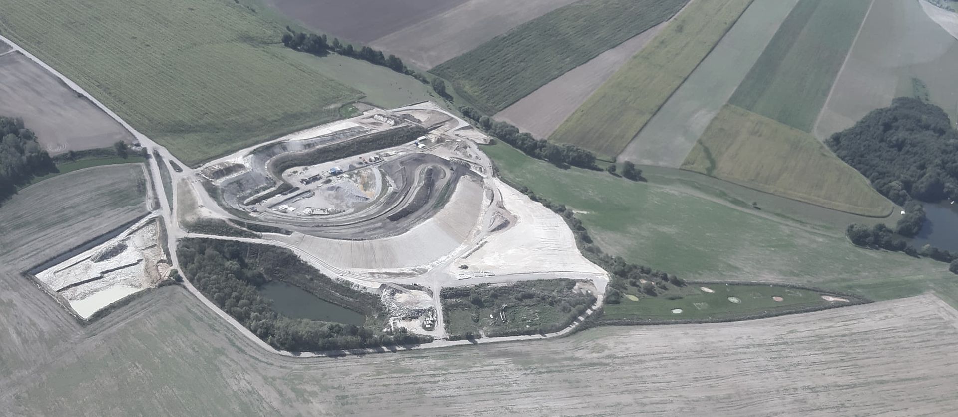 Luftaufnahme einer von grünen Feldern umgebenen Baustelle mit Erdarbeiten, einem Teich und in Betrieb befindlichen Maschinen.