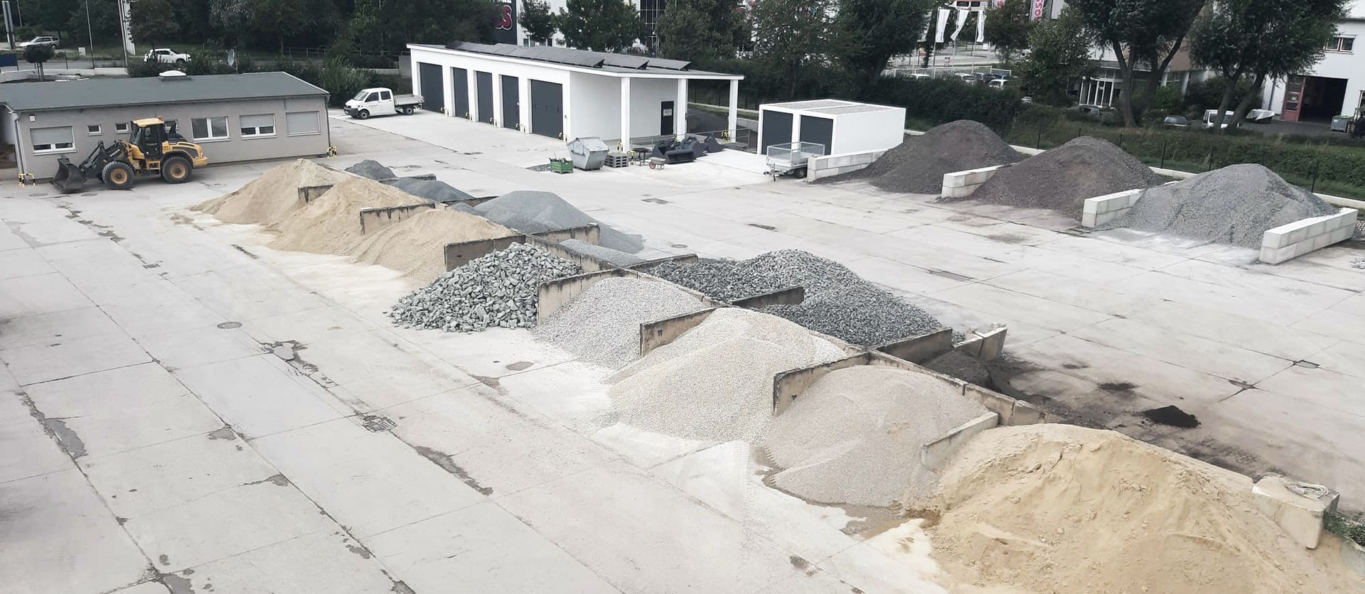Luftaufnahme eines Bauhofs mit Stapeln von verschiedenen Kies- und Sandmaterialien und einem Ladefahrzeug im Vordergrund.