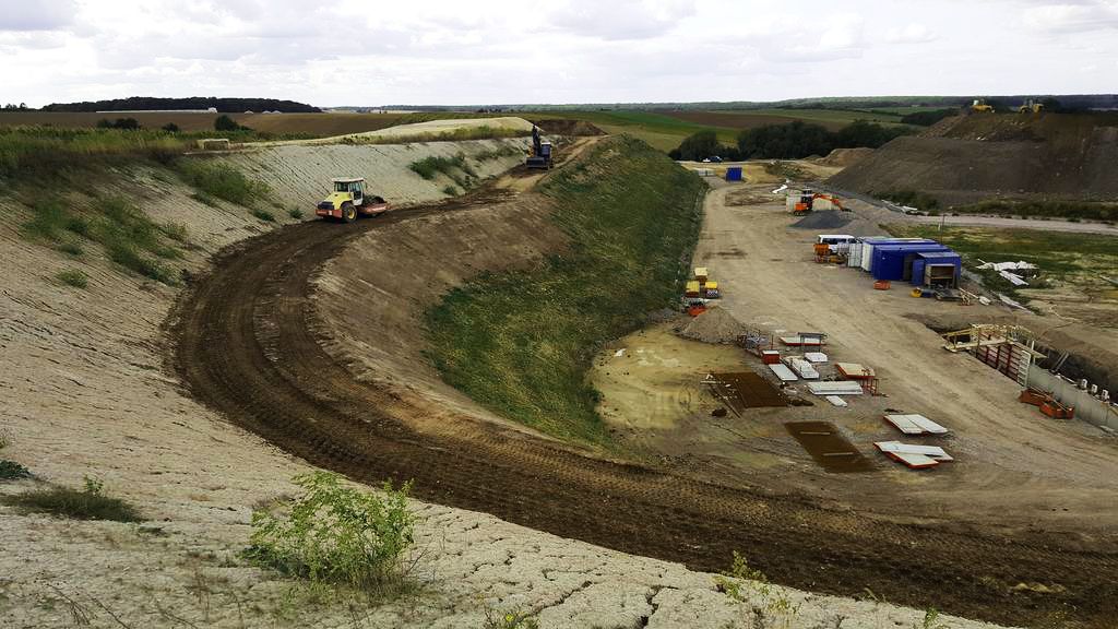 Eine Baustelle mit Maschinen, einem geschwungenen Feldweg und Lagercontainern in einer ländlichen Landschaft unter bewölktem Himmel.