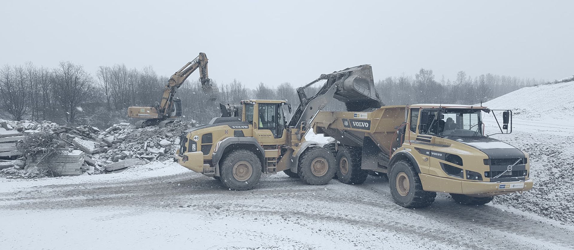 Zwei Volvo-Baufahrzeuge bei der Arbeit im Schnee: Eines lädt Schutt auf, während das andere Material auf einer Baustelle abtransportiert.
