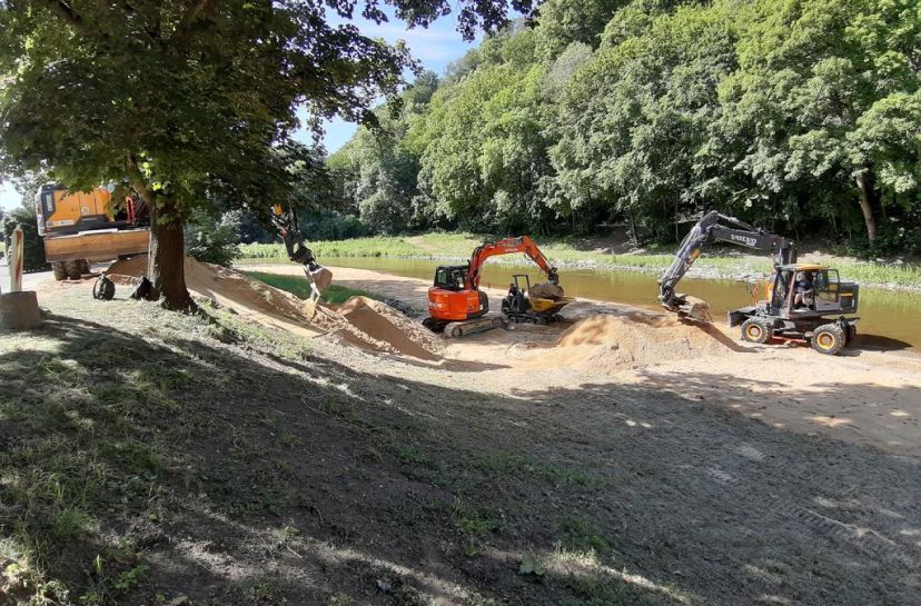 Bagger und ein Muldenkipper arbeiten an einem sandigen Flussufer inmitten von üppigem Grün, um das Gebiet für die künftige Entwicklung vorzubereiten.