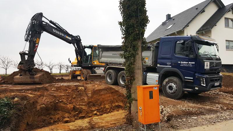 Baustelle mit einem Volvo-Bagger, der neben einem großen Muldenkipper Erde aushebt, mit einem Haus und einem Stromkasten im Hintergrund.
