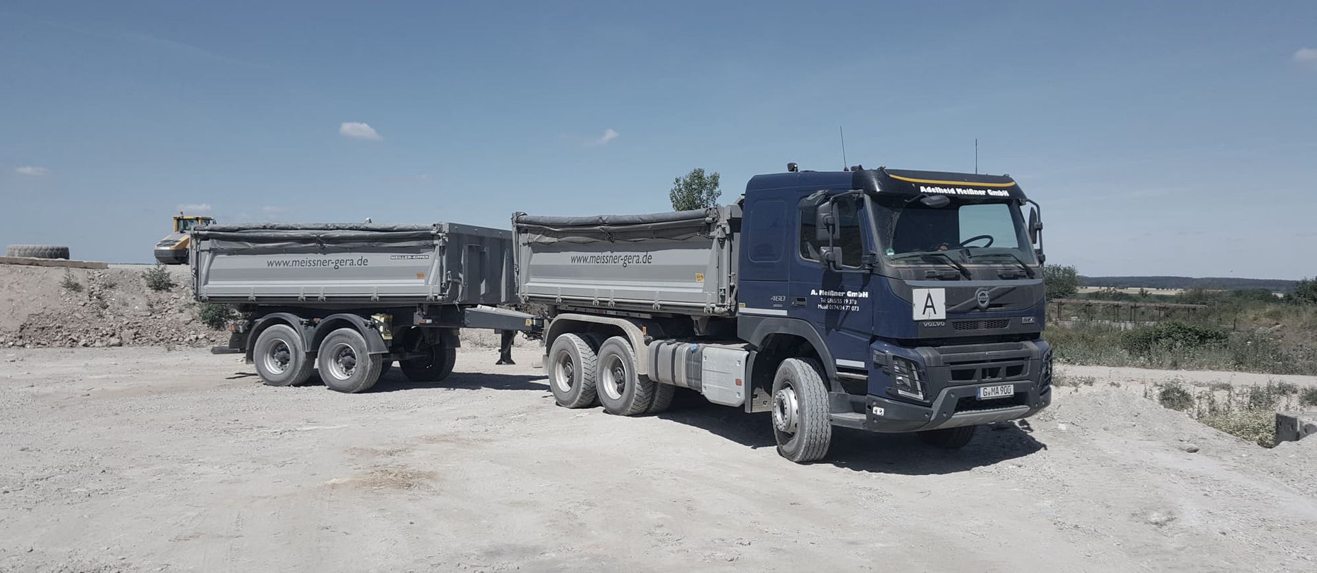 Ein blauer Volvo-Muldenkipper mit zwei silbernen Anhängern parkt auf einer staubigen Baustelle unter freiem Himmel.