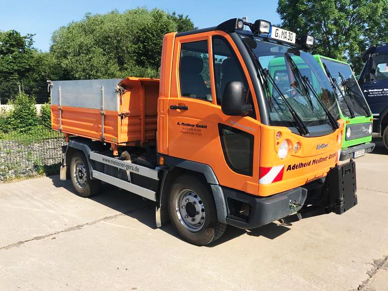 Ein orangefarbener Nutzfahrzeug mit Metallkippmulde, geparkt im Sonnenlicht, mit Bäumen im Hintergrund, die sein industrielles Design zur Geltung bringen.